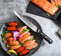 Een Paudin mes met damaststalen lemmet ligt op een bord met zalmsashimi en viseieren. Gekookte zalmfilet ligt ernaast.