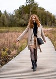 Roodharige vrouw met beige jas, zwarte coltrui, rok en laarzen, met zwarte schoudertas.