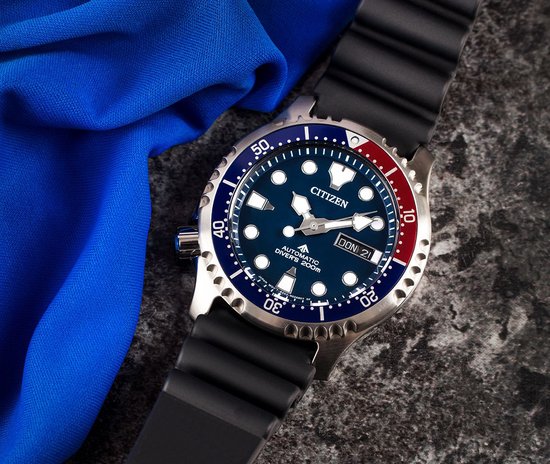 Citizen Promaster horloge met blauwe wijzerplaat, rood-blauwe lunette en datumweergave, naast een blauwe doek.