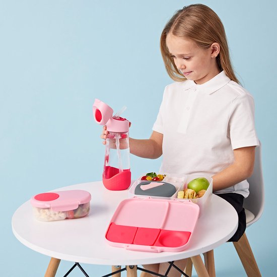 Fille aux cheveux blonds en chemise blanche boit dans une gourde b.box rose à côté d'une boîte à lunch rose ouverte.