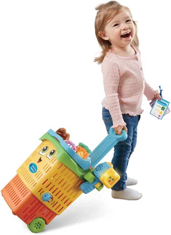 Blond meisje in roze trui, trekt een felgekleurde winkelwagen met speelgoedboodschappen.