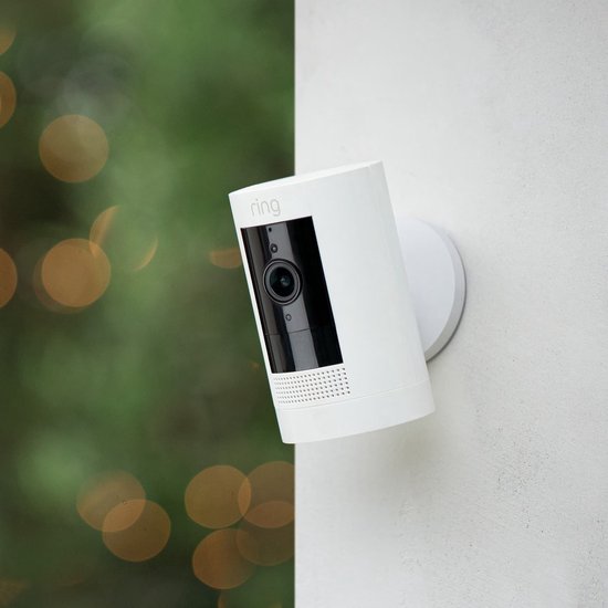 Witte Ring Stick Up Cam bevestigd aan een muur, met groene bokeh op achtergrond.
