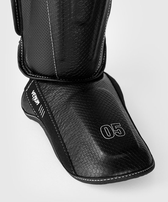 Zwarte scheenbeschermer met schubbenpatroon, witte stiksels en '05' op de voet.