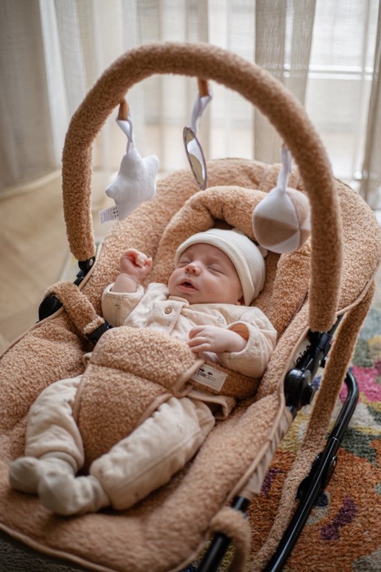 Baby in beige outfit en mutsje slaapt in een zachte bruine wipstoel met speelgoed.