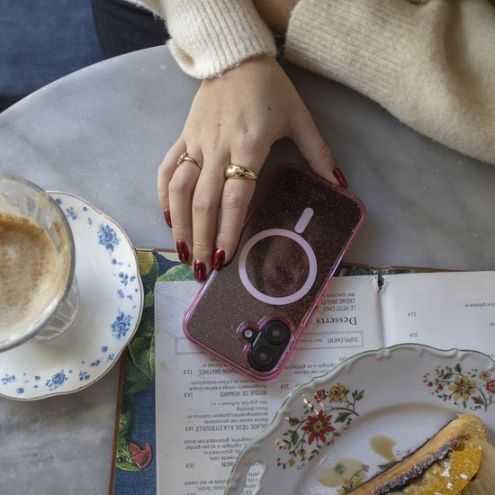 Roze glitter telefoonhoesje met MagSafe ring, in een hand met rode nagels, naast een kop koffie en dessert.