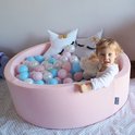 Fille dans un parc à balles KiddyMoon rose pâle avec des balles bleues, blanches et roses. Deux coussins étoiles et un ours en peluche.