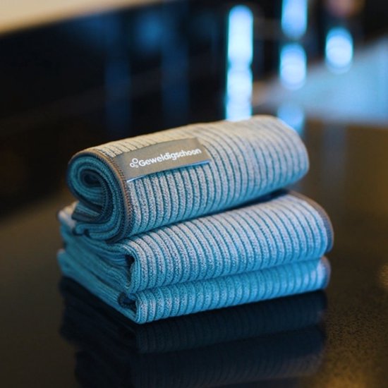 Trois chiffons en microfibre bleu-gris pliés sur une surface noire. Le chiffon supérieur a une étiquette indiquant 'Geweldigschoon'.
