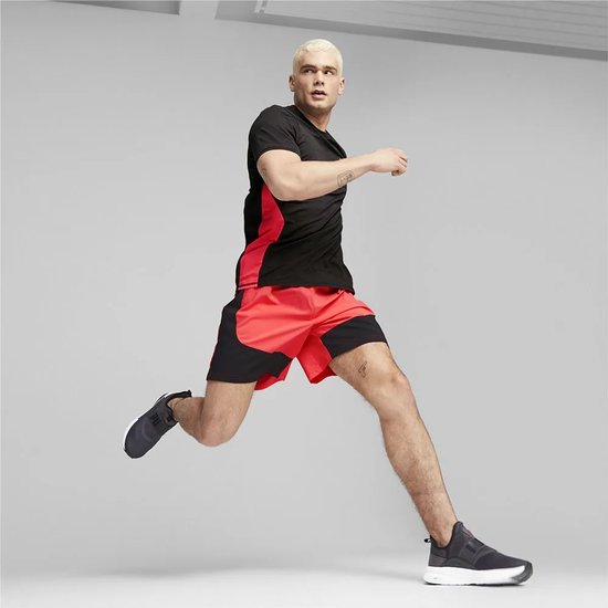 Man in zwart shirt met rode accenten, rode shorts en donkergrijze schoenen, in de lucht springend.