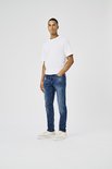 Man in donkerblauwe jeans, wit t-shirt en beige sneakers, van de zijkant gezien.