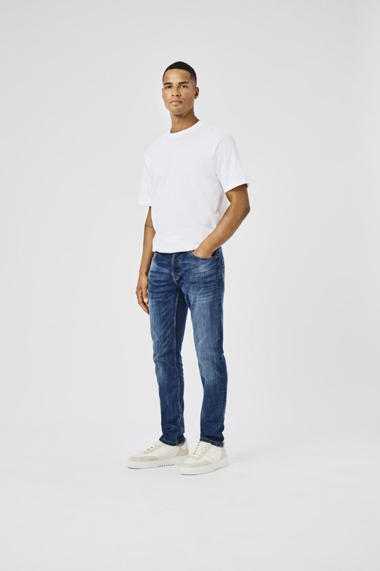 Man in donkerblauwe jeans, wit t-shirt en beige sneakers, van de zijkant gezien.