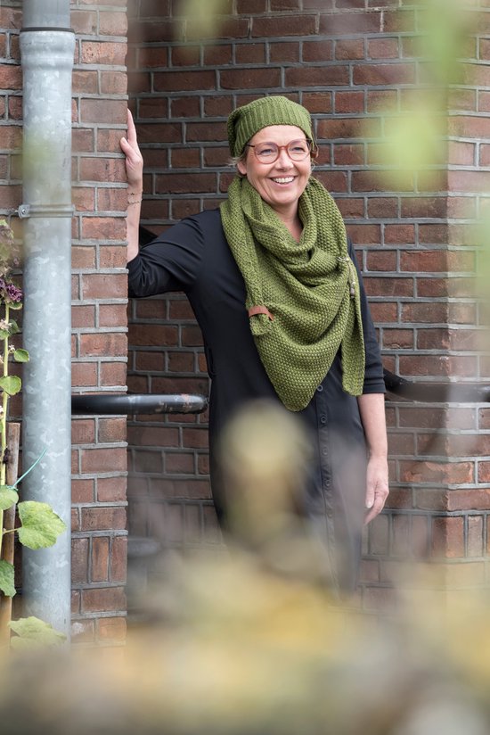 Een lachende vrouw met bril in een gebreide groene muts en een grote sjaal, leunend tegen een bakstenen muur.