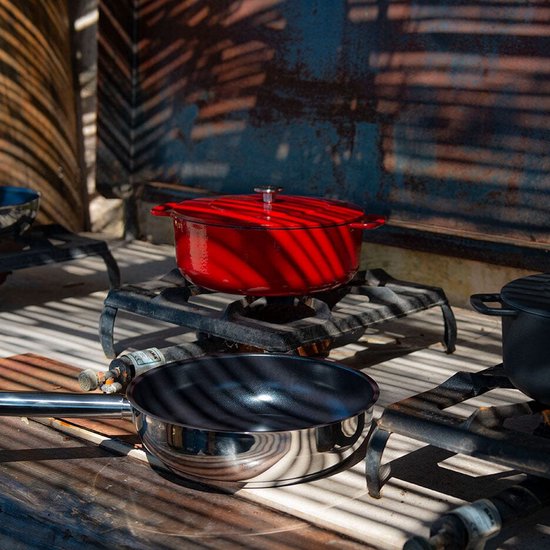 Rode gietijzeren braadpan en een roestvrijstalen koekenpan op gasbranders, met schaduwen van palmbladeren.