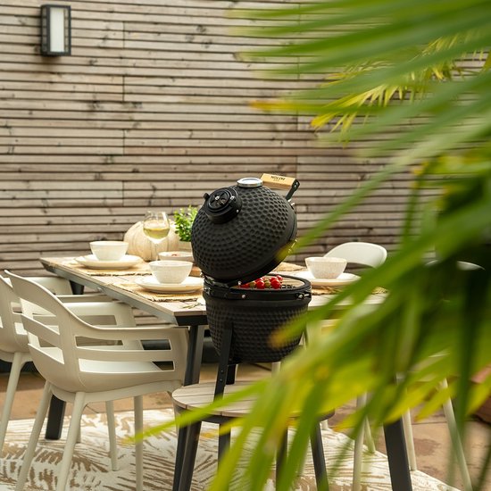 Zwarte kamado barbecue met open deksel op een tuintafel, tussen planten en stoelen.
