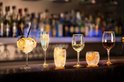 Zes glazen op een bar, gevuld met drank en fruit, waaronder citroen, sinaasappel en bessen.