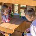 Twee jonge meisjes spelen aan een houten speelhuisje met een aanrecht en gootsteen.