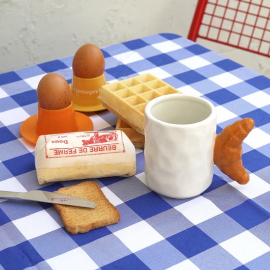 Witte mok met croissant-oor op een blauw-wit geblokte tafel, naast eieren, boter en toast.