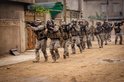 Militairen in camouflageuniformen lopen in linie op een stoffige straat, wapens in de aanslag.