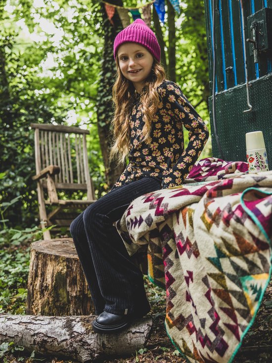 Lachend meisje met blond haar in knot, draagt een zwart shirt met bloemenprint en fuchsia corduroy broek.