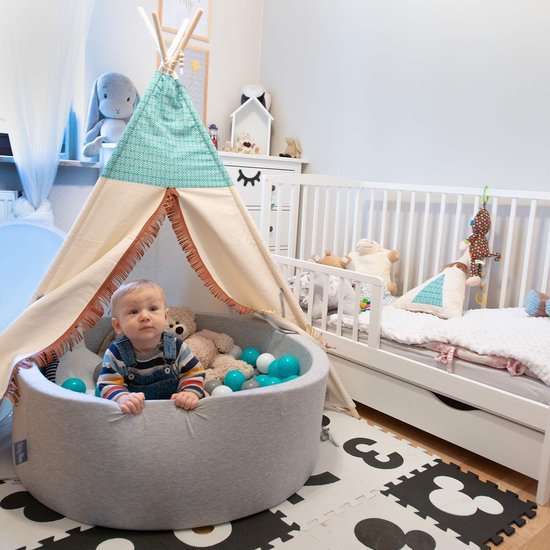 Un bébé est assis dans une piscine à balles grise près d'une tente tipi, à côté d'un lit d'enfant blanc.