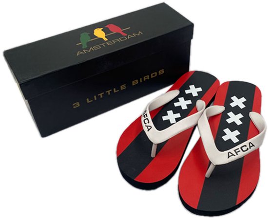 Slippers met rode en zwarte banen en drie Andreaskruizen, naast een zwarte doos met '3 LITTLE BIRDS AMSTERDAM'.