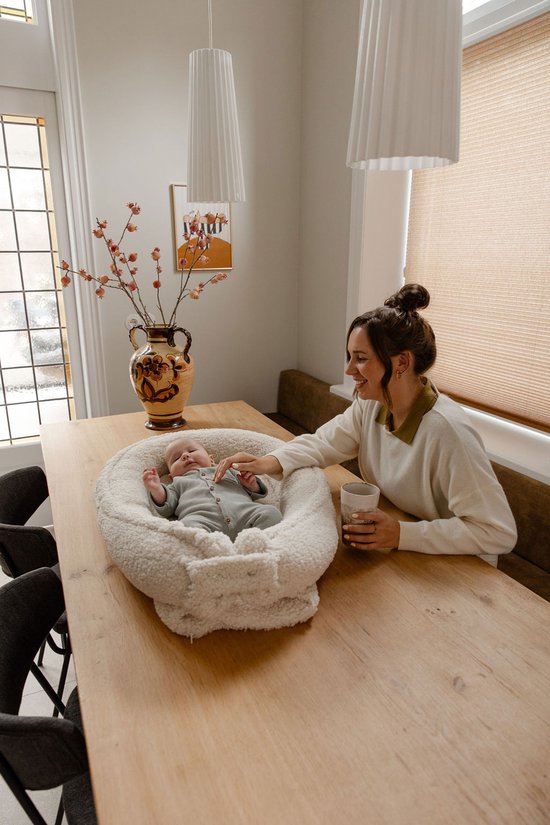 Een vrouw zit aan tafel en praat met een baby in een wit teddy babynestje voor haar.