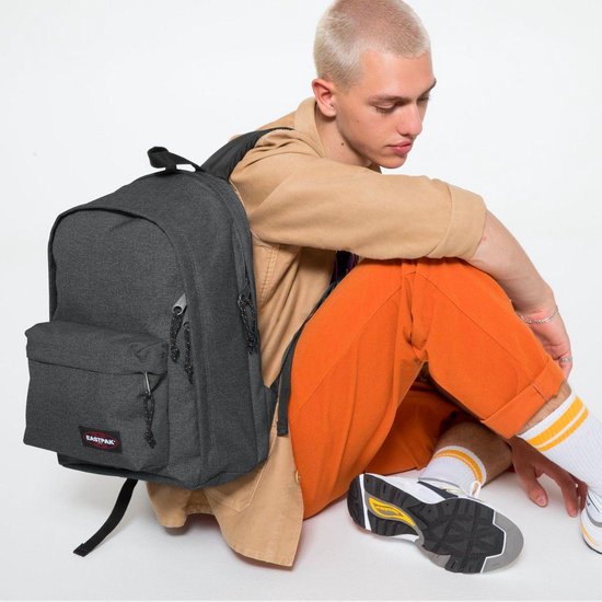 Model zittend naast een donkergrijze Eastpak rugzak, draagt oranje broek.
