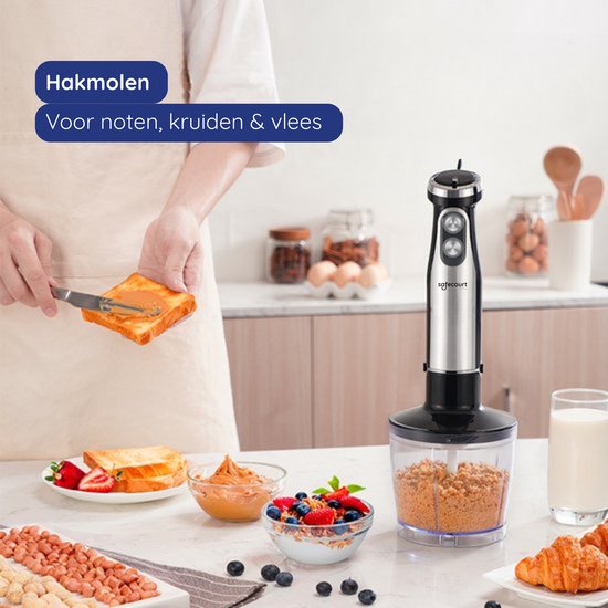 Zilverkleurige staafmixer met zwarte basis en hakmolen. Naast een glas melk en een croissant. "Hakmolen. Voor noten, kruiden & vlees."