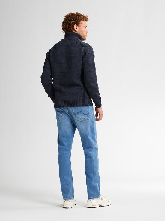 Man met krullend rood haar, draagt een donkerblauwe gebreide trui en spijkerbroek.