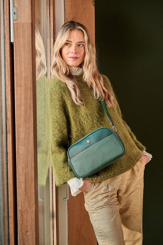 Vrouw met blonde haren in een groene trui en glimmende broek draagt een groene crossbody tas.