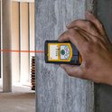 Een hand houdt een gele en zwarte afstandsmeter vast, een laserlijn wordt geprojecteerd over een bouwterrein.