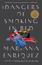 Boekcover: "The Dangers of Smoking in Bed" door Mariana Enriquez. Een vrouw met rood haar ligt op bed met een blauwe deken. "Extraordinary—and extraordinarily ominous." - OPRAH DAILY