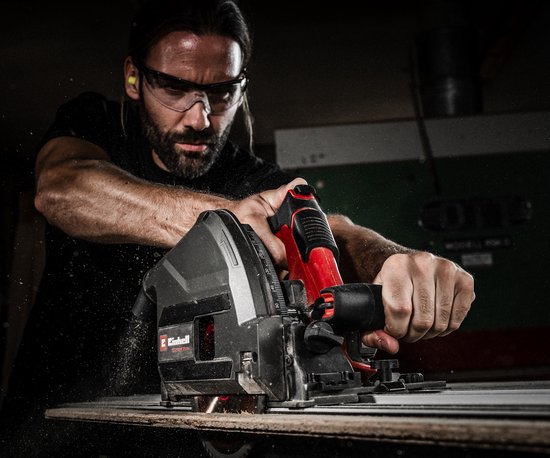 Man met veiligheidsbril gebruikt de Einhell invalzaag op een houten plank, met rondvliegend zaagsel.