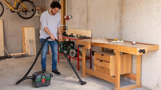 Man met grijze trui stofzuigt werkplaats met groene en zwarte ketelstofzuiger.