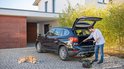 Man met krultang stofzuigt kofferbak zwarte auto. Ketelstofzuiger met groene en rode accenten op oprit.