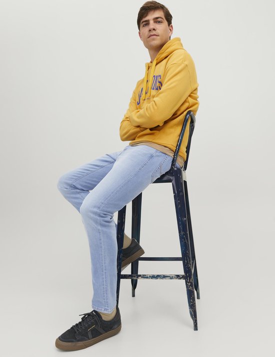 Model zit op een kruk, draagt een gele Jack & Jones hoodie en lichtblauwe slim fit jeans.