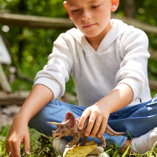 Jongen met witte hoodie speelt met bruine dinosaurus met rode accenten op gras.