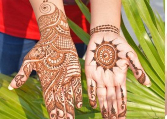 Twee handen met gedetailleerde henna-tatoeages. De linkerhand heeft een complex, omvangrijk ontwerp. De rechterhand heeft een zonmotief op de palm en stippen op de vingers.