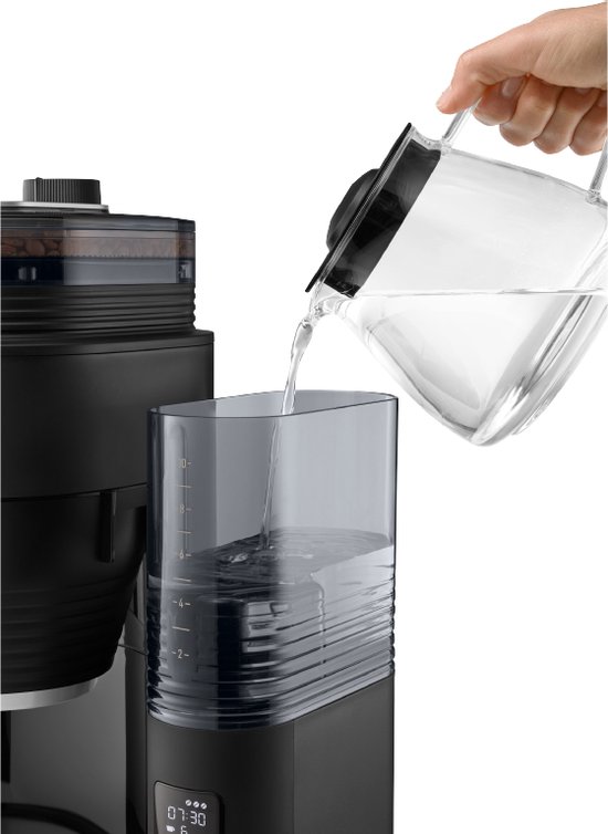 Een hand giet water uit een glazen kan in het waterreservoir van een zwart koffiezetapparaat.