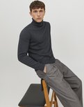Man zittend op een stoel, draagt een donkergrijze coltrui en een grijze pantalon met krijtstrepen.