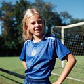 Jong meisje met blonde paardenstaart, beugel en schelpenketting, lachend in een blauw Chelsea FC voetbalshirt met witte accenten.