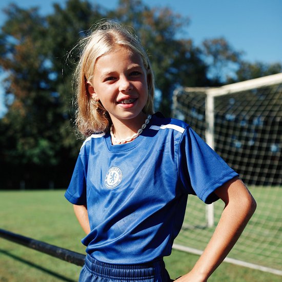 Jong meisje met blonde paardenstaart, beugel en schelpenketting, lachend in een blauw Chelsea FC voetbalshirt met witte accenten.