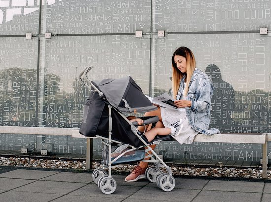 Vrouw in spijkerjasje leest bij een grijze buggy met kind.