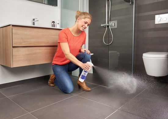 Een vrouw met een oranje shirt en spijkerbroek die Secucare antislip spray op de badkamervloer spuit.