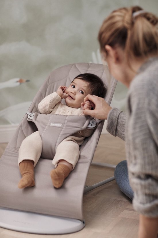 Baby in zandgrijs wipstoeltje, kijkt naar persoon.