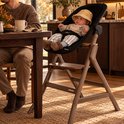 Baby in hoedje zittend in een zwarte Deryan bouncer bevestigd op een donkerbruin houten kinderstoelframe.