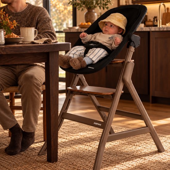 Baby in hoedje zittend in een zwarte Deryan bouncer bevestigd op een donkerbruin houten kinderstoelframe.