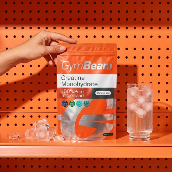 Hand toont een GymBeam Creatine Monohydraat zak, niet-gearomatiseerd, naast een glas water en ijs.