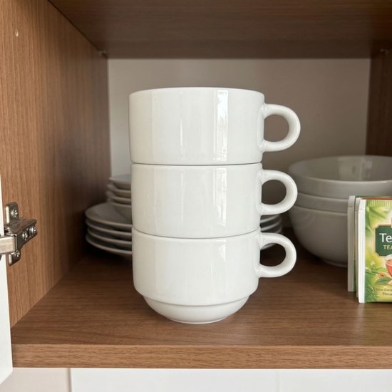 Drie witte stapelbare mokken, opgestapeld in een keukenkastje naast theezakjes.