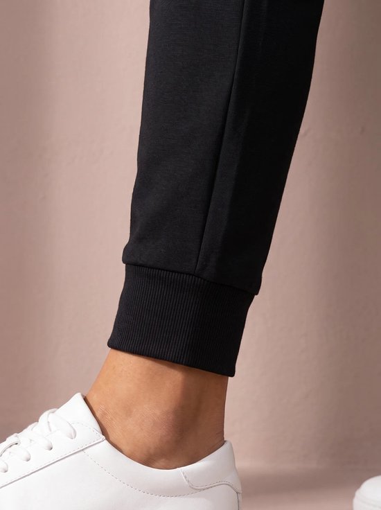 Close-up van de elastische enkelband van een zwarte sportbroek, gedragen door een model met witte sneakers.