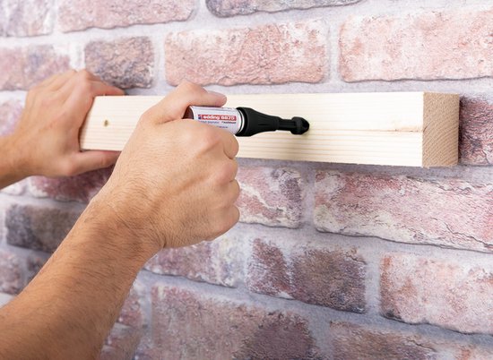Handen markeren een houten balk met de Edding 8875 deep hole spray marker op een bakstenen muur.